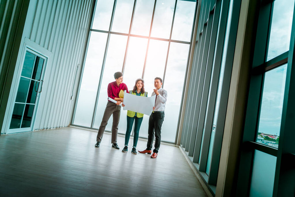 Три инженера строительной команды и дизайнер интерьеров проводят мозговой штурм в пустом помещении с планом проекта. - Фото, изображение