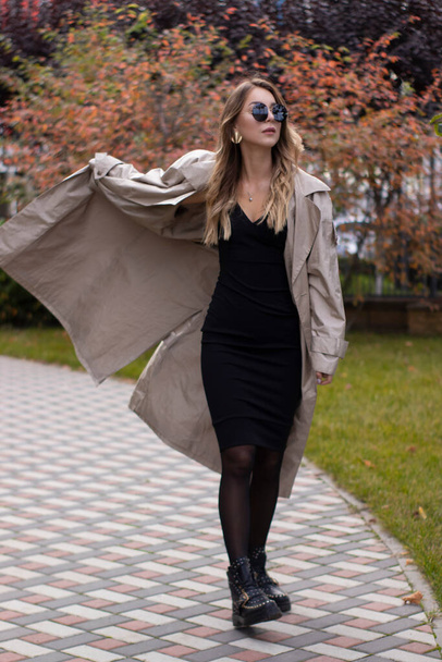 atrakcyjna szczupła młoda brunetka w modnym okopie, czarnej sukience i okularach przeciwsłonecznych w parku. jesień, liście żółte - Zdjęcie, obraz