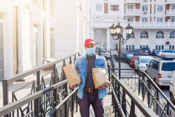 Entrega hombre sosteniendo bolsa de papel con comida en la entrada blanca del fondo de la casa, hombre de entrega de alimentos en máscara protectora - Foto, imagen