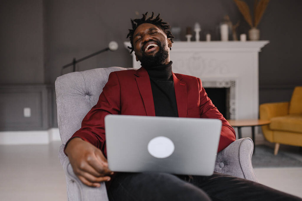 Un bel ragazzo afroamericano sorridente seduto a guardare lo schermo del computer. Uomo millenario focalizzato che lavora al progetto in ufficio moderno o studia online a casa. - Foto, immagini