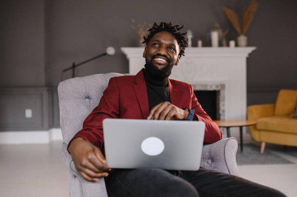 Smiling handsome young african american guy sitting, looking at computer screen. Focused millennial biracial man working on project in modern office or studying online at home. - Foto, Imagen