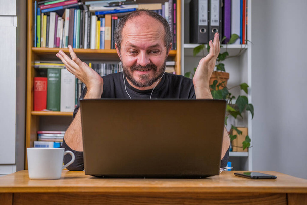 Człowiek pracujący online z domu na laptopie komputerowym za biurkiem vintage, ze słuchawkami, mówiący i wyjaśniający kolegom na spotkaniu online, gesty ramion - Zdjęcie, obraz
