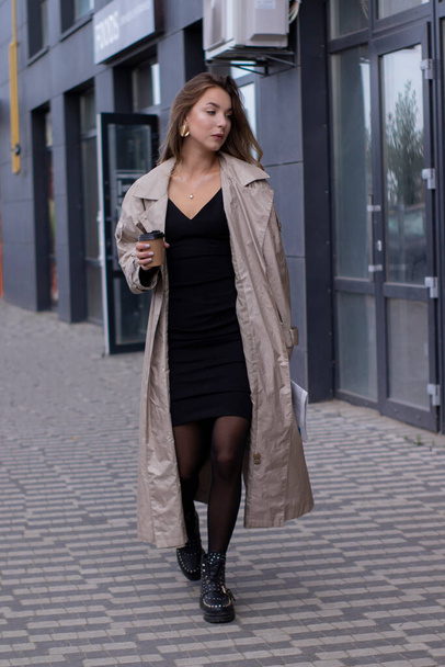beautiful young brunette in fashionable trench coat and black dress walking through the street. autumn, yellow leaves. - Photo, Image