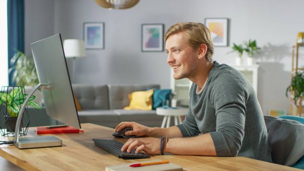 Portrait of the Handsome Smiling Man Working on Personal Computer while Sitting at His Desk. In the Background Stylish Cozy Living Room. Young Freelancer Working From Home. - Photo, Image
