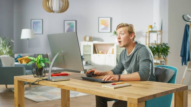 Portrait of Surprised Handsome Man Working on Personal Computer Has Stroke of Luck, Emotionally Showing it. In the Background Stylish Cozy Living Room. - 写真・画像