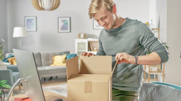 Handsome Young Man in Living Room Opening Cardboard Box Package With Interest. - Fotoğraf, Görsel