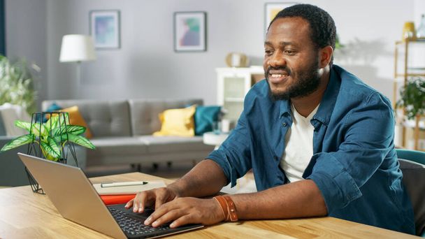 Handsome Smiling Black Man Using Laptop While Sitting at the Desk of His Cozy Living Room. - Foto, immagini