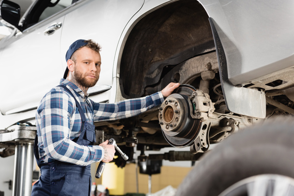 technician looking at camera while standing near lifted automobile with wrench on blurred foreground - Foto, Imagen