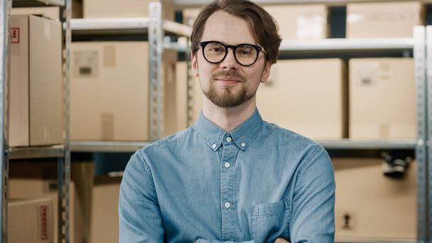 Portrait of the Handsome Warehouse Inventory Manager Standing with Crossed Arms and Smiling. Smart Man Wearing Glasses with Rows of Shelves Full of Cardboard Boxes and Parcels Ready for Shipment. - 写真・画像