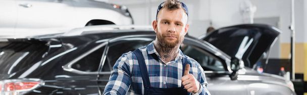 young mechanic showing thumb up while looking at camera in workshop, banner - Фото, изображение