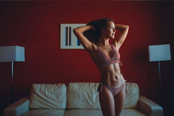 a young girl in a striped swimsuit is engaged in stretching and balance in a red room room with furniture and floor lamps and a painting. - Fotó, kép