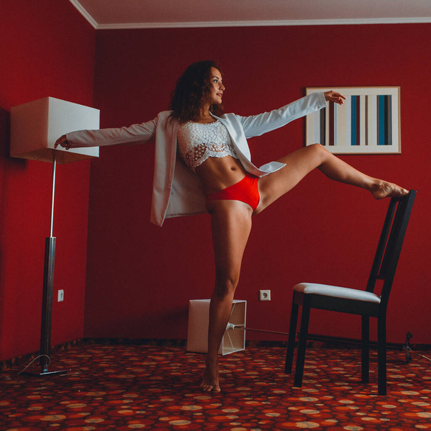 fiatal lány fehér kabátban és piros fürdőnadrágban, részt vesz nyújtás és az egyensúly egy piros szobában egy szobában, bútorokkal és padló lámpák - Fotó, kép