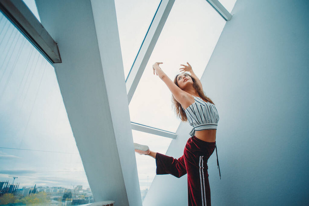 a young girl in sweatpants and a tank top is engaged in stretching in a bright room near a panoramic window - 写真・画像