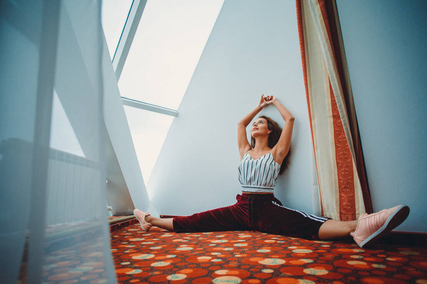 a young girl in sweatpants and a tank top is engaged in stretching in a bright room near a panoramic window - Photo, Image