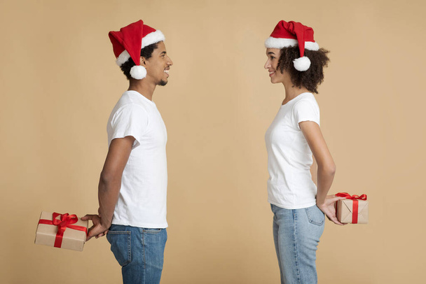 Sorridente giovane uomo e donna in cappelli di Babbo Natale si guardano l'un l'altro e tengono regali a sorpresa dietro la schiena - Foto, immagini