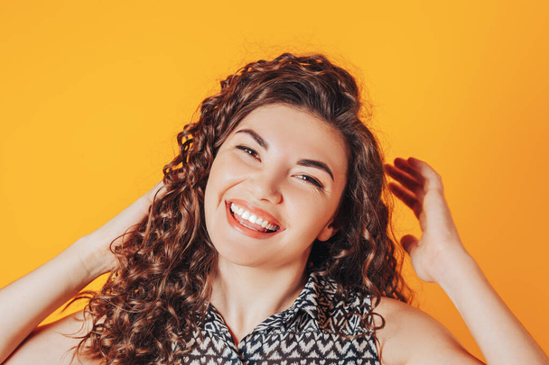 Beautiful woman with curly hair posing at camera on yellow background, hands up. - Foto, imagen
