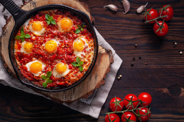Fried eggs with tomatoes and vegetables. Shakshuka in a cast iron portioned pan on a wooden background top view. Traditional breakfast dish. - Photo, image