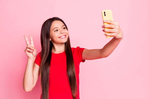 Photo of lovely sweet little lady long hairstyle raise hand hold telephone show v-sign take selfie video message look screen wear casual red t-shirt isolated pink color background - 写真・画像