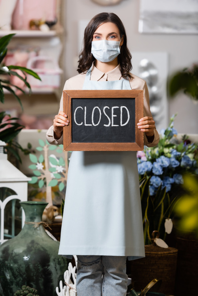 young florist in medical mask holding board with closed lettering in flower shop on blurred foreground - Photo, Image