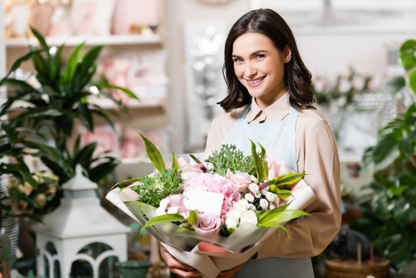 young florist smiling at camera while holding bouquet with empty tag near plants on blurred background - Photo, Image