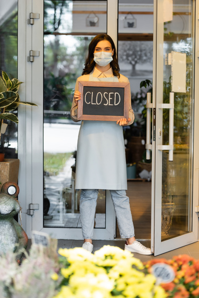 young florist holding board with closed lettering near entrance of flower shop and flowers on blurred foreground - Фото, изображение