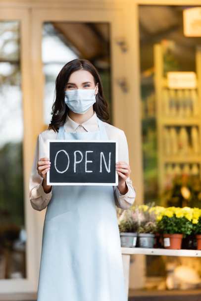 positive florist in protective mask holding board with open lettering near flower shop on blurred background - Foto, immagini