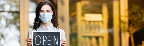 positive florist in medical mask holding board with open lettering near flower shop on blurred background, banner - Photo, Image
