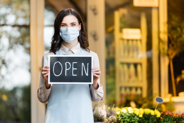 cheerful florist in protective mask holding board with open lettering near flower shop on blurred background - Foto, immagini