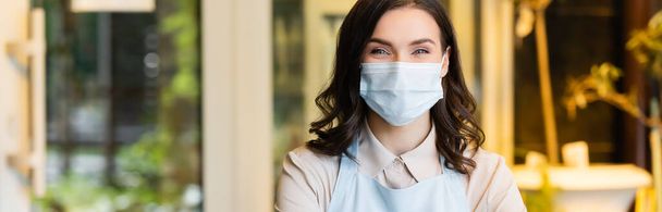 positive florist in medical mask looking at camera in flower shop, banner - Photo, Image