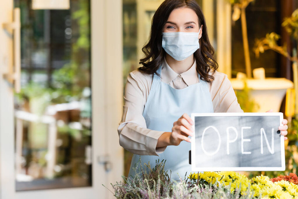 happy florist in medical mask holding board with open lettering near flower shop on blurred background - Photo, Image