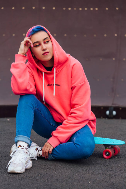 Portrait of a trendy pretty young girl dressed in pink hoodie and jeans sitting next to the skateboard court on her blue plastic skateboard. - Foto, immagini