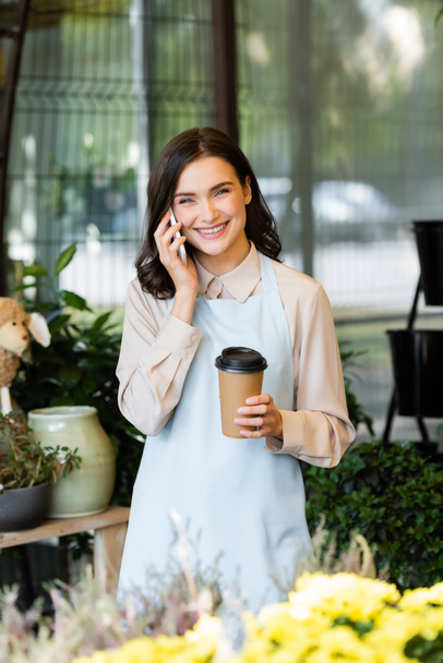 cheerful florist talking on smartphone and holding coffee to go near blurred flowers on foreground  - Photo, Image