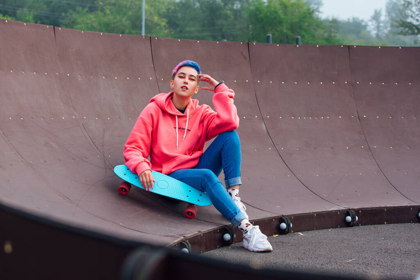 Stylish young woman with short colored hair sitting with her blue plastic skateboard in skatepark. Youth concept. - Photo, image