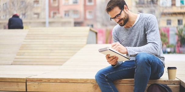 Crop young handsome male writer in casual wear and eyeglasses sitting on wooden step on city street while taking notes in planner for preparing new article - Фото, изображение