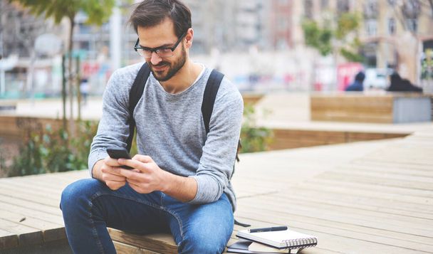 Positive male entrepreneur using cellphone and sitting on wooden bench near notebooks while working online on new startup concept outside - Photo, image