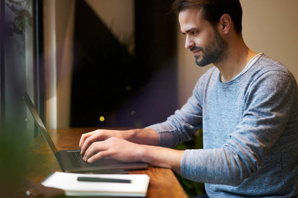 Side view of bearded male freelancer in casual style clothing working on laptop while sitting at wooden table with notepad and pen - Φωτογραφία, εικόνα