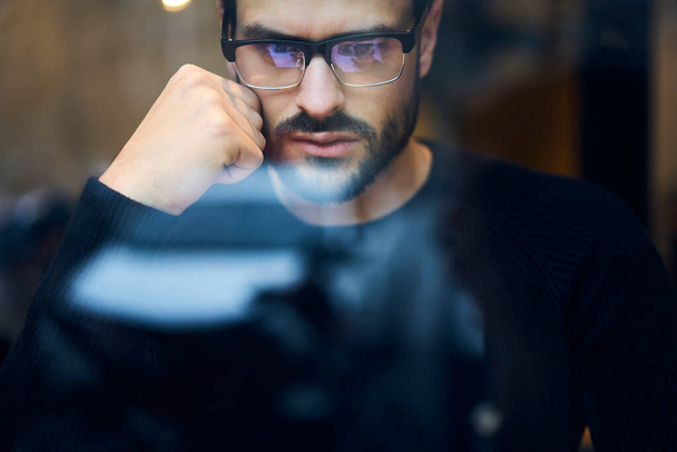 Keresztül üveg kilátás a termés komoly fókuszált szakállas férfi szemüvegben és alkalmi sötét pulóver támaszkodik a kezét, és fontolgatja - Fotó, kép