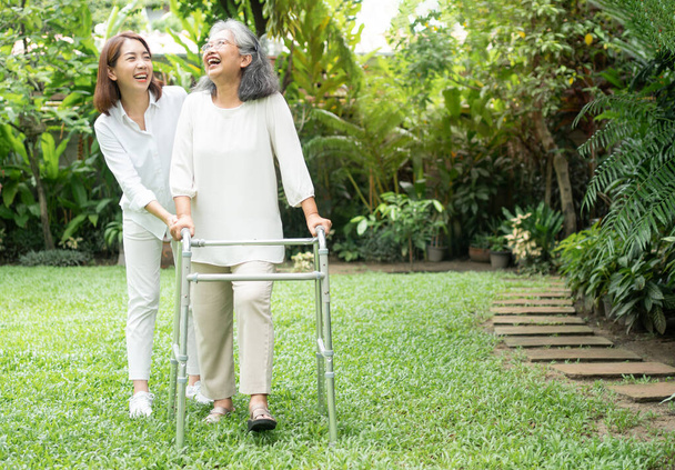 Een oude Aziatische vrouw gebruikt een looprek en loopt in de achtertuin met haar dochter. Concept van gelukkig pensioen Met zorg van een zorgverlener en spaargeld en senior ziektekostenverzekering - Foto, afbeelding