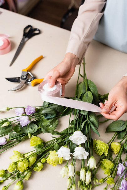 vista cortada de florista segurando fita decorativa perto de flores eustoma, tesouras e tesouras na mesa - Foto, Imagem