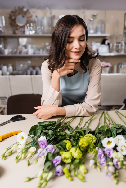 happy florist looking at eustoma flowers on table in flower shop on blurred foreground - Fotó, kép