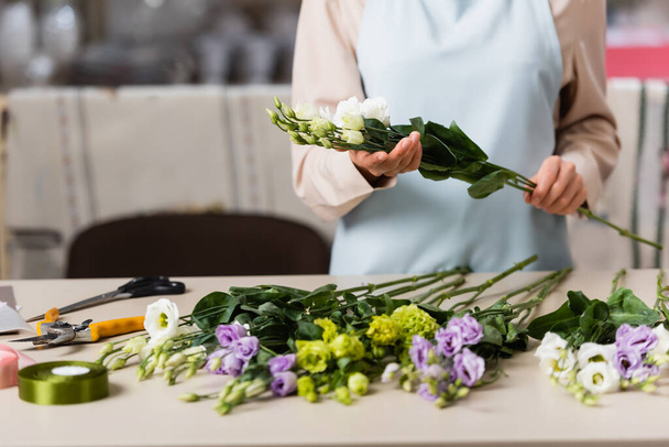 vista parcial de florista sosteniendo flores de eustoma mientras hace ramo en la tienda de flores en primer plano borroso - Foto, Imagen