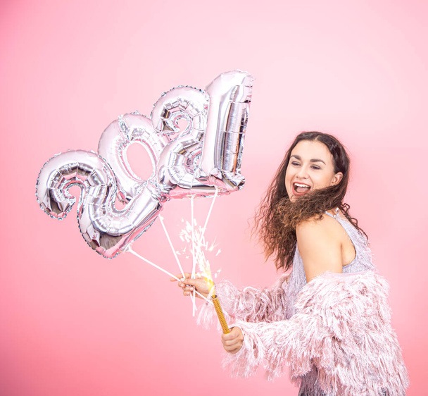 Vrolijk feest meisje brunette met krullend haar feestelijk gekleed met een vuurwerk kaars in haar hand en zilveren ballonnen in van de nummers 2021 op een roze achtergrond, concept van het nieuwe jaar - Foto, afbeelding