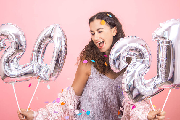 Krásná brunetka strana dívka s kudrnaté vlasy a slavnostní oblečení pózovat na růžové pozadí studia s konfety a drží stříbrné balónky z čísel 2021 v ruce, šťastný nový rok koncept - Fotografie, Obrázek