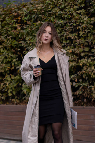 beautiful slim young brunette in fashionable trench coat and black dress with a paper cup coffee - Foto, Imagem