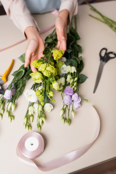 top view of florist holding eustoma flowers near scissors and decorative tape while making bouquet in flower shop - Photo, Image