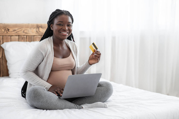 Online nakupování. Šťastná těhotná žena s notebookem a kreditní kartou na posteli - Fotografie, Obrázek