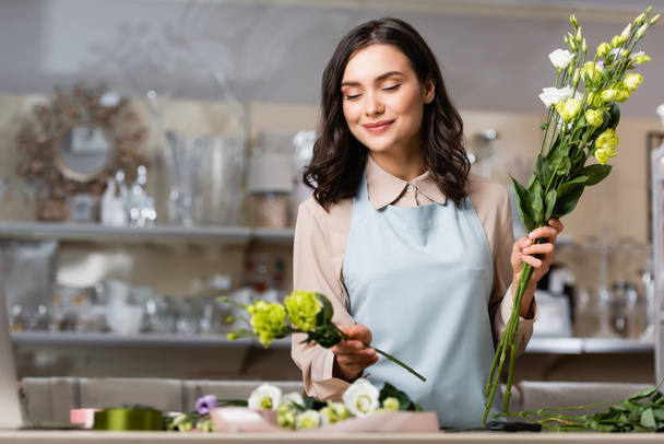 smiling florist in flower shop holding eustoma flowers while arranging bouquet on blurred foreground - Photo, Image
