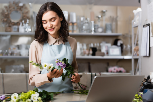 smiling florist holding eustoma flowers near laptop on blurred foreground - Foto, afbeelding