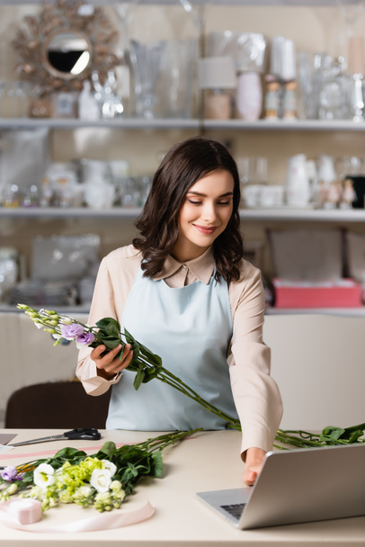 улыбающийся флорист, держащий цветы эустомы при использовании ноутбука возле стойки с вазами на размытом фоне - Фото, изображение