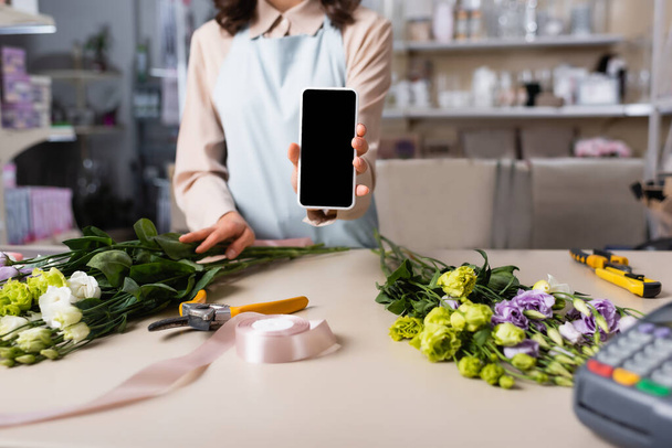 Ausgeschnittene Ansicht des Blumenhändlers zeigt Smartphone mit leerem Bildschirm mit Eustoma-Blumen auf Schreibtisch auf verschwommenem Hintergrund - Foto, Bild